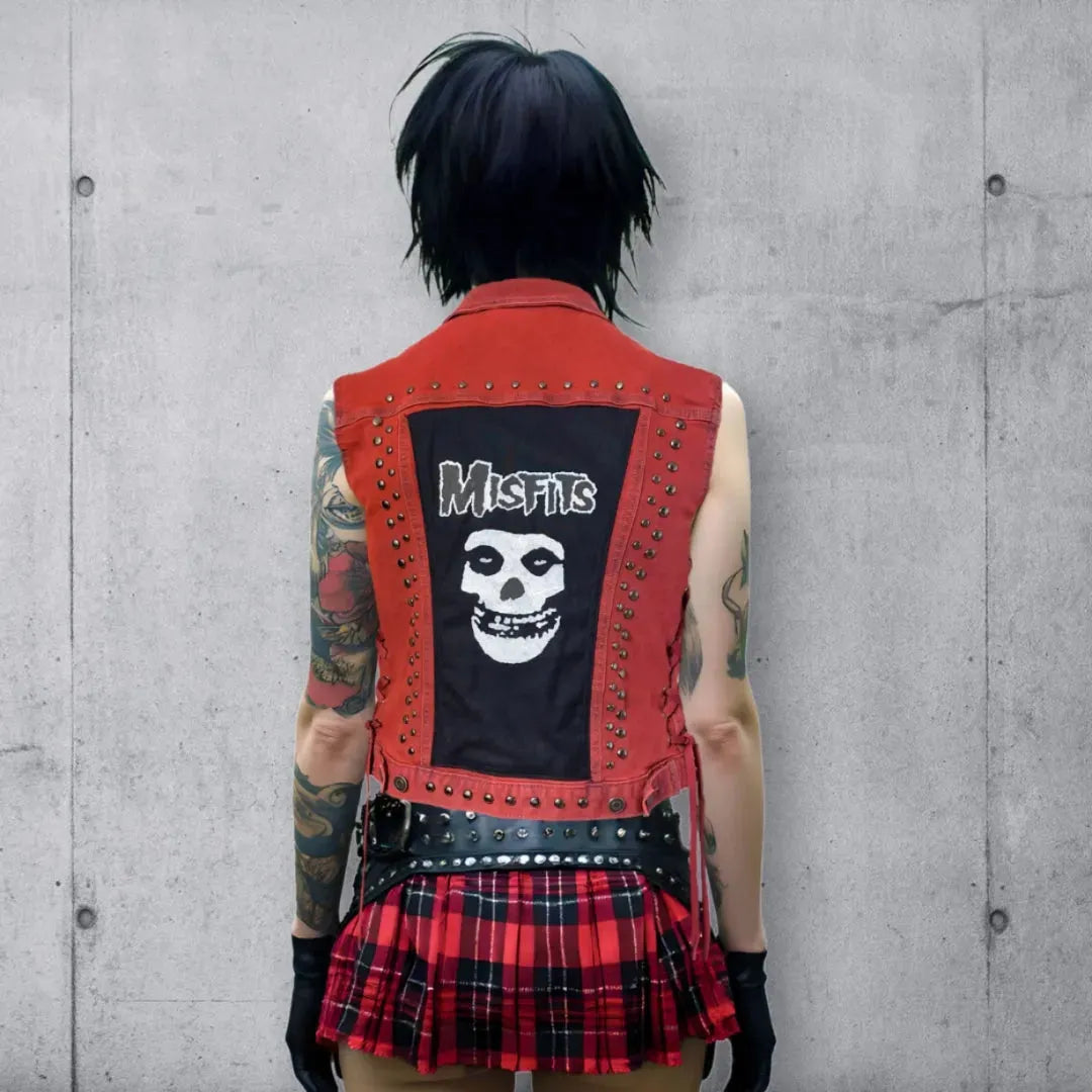 Back view of a woman in a red vest with a Misfits crimson ghost face and band name above it, round black punk studs, a red plaid mini skirt, and tattoos, standing against a concrete wall backdrop.