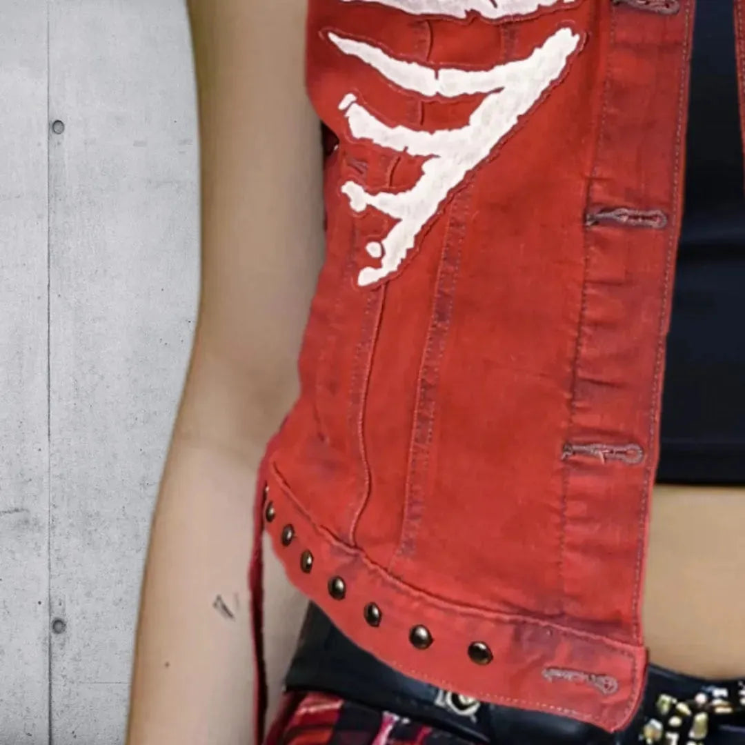 Lower right side detail of a red vest with white skeleton rib cage patches, the jacket hem lined with gunmetal round punk studs, worn over a black crop top and red plaid mini skirt.