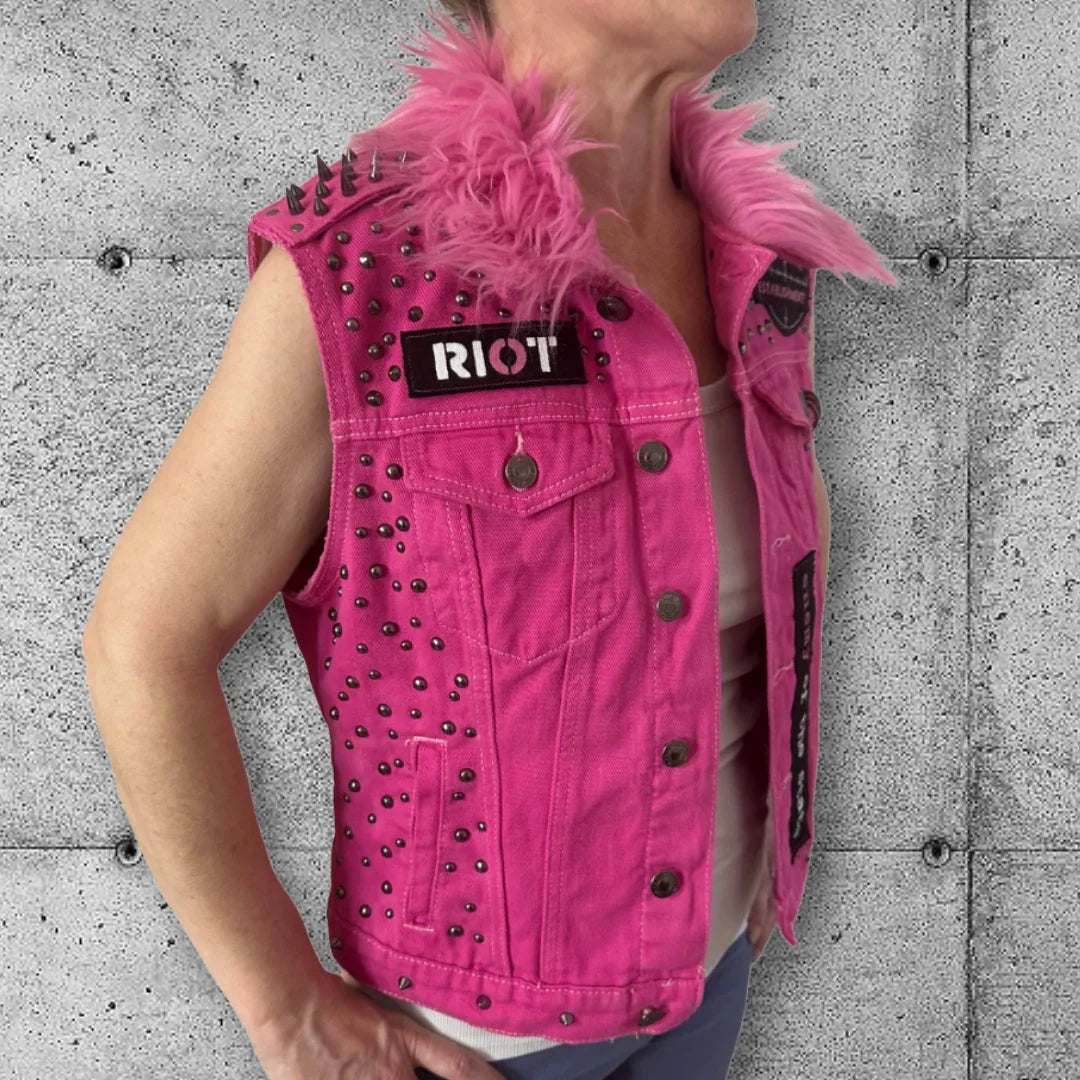 Woman wearing a pink punk vest with metal studs, spikes, faux fur collar, and RIOT patch.
