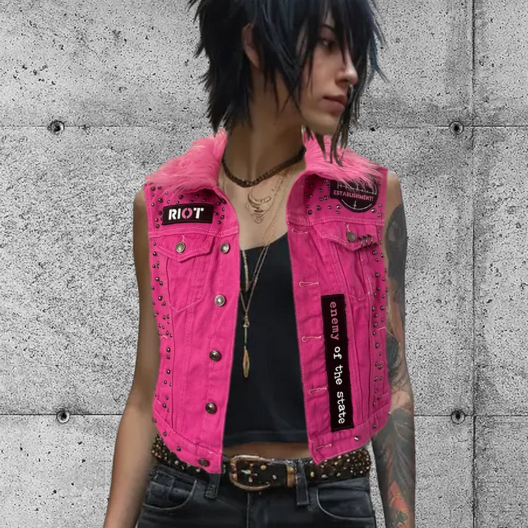 Woman in punk style pink studded vest with black patches saying 'riot' and 'enemy of the state'.