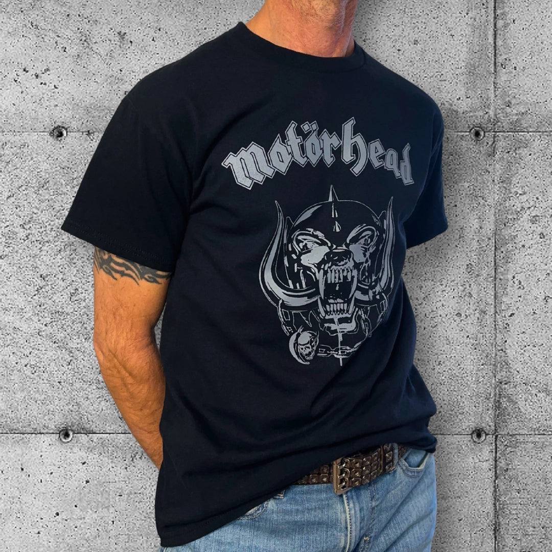 Man wearing black Motörhead logo punk t-shirt and jeans, standing by a concrete wall.