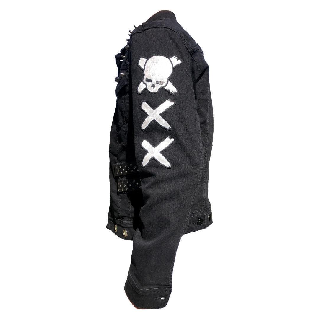 Left side view of black punk-style denim jacket featuring skull and cross bones designed patches, black leather straps with metal studs, and two white painted 'X'  on the sleeve. Perfect for alternative fashion enthusiasts.